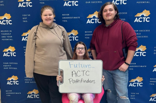 Three high school students smiling in front of an ACTC back wall. Two are standing and one is sitting holding a white board that says "Future ACTC Pathfinders"