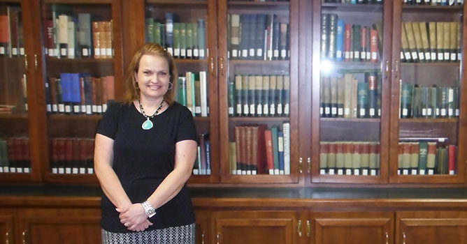 Assistant Professor of Mathematics and advisor, Cindy Shelton stands in front large oak bookcase in the meeting room at the Masbach Memorial Library.