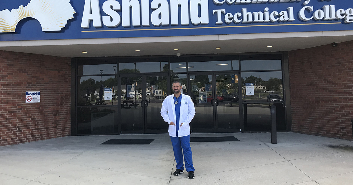 A man in blue scrubs and a white lab coat smiling