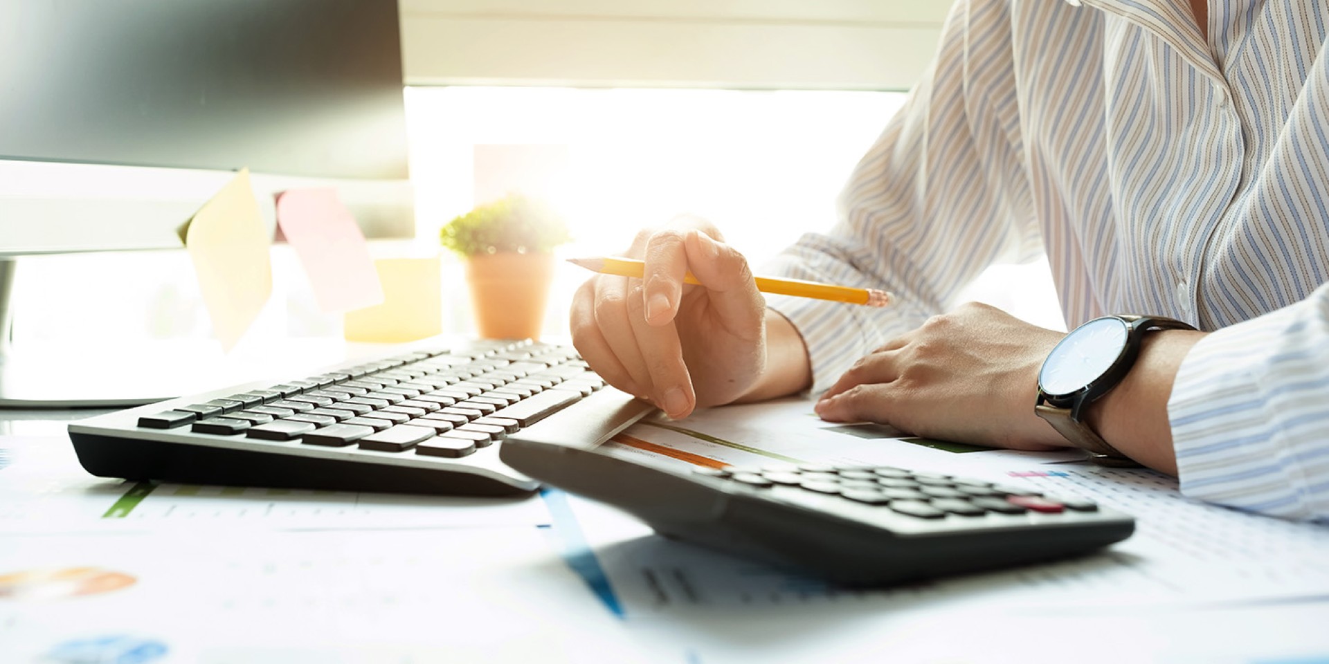 The chest and hands of someone who is sitting in front of a computer, a calculator, and some paper forms while holding a pencil.