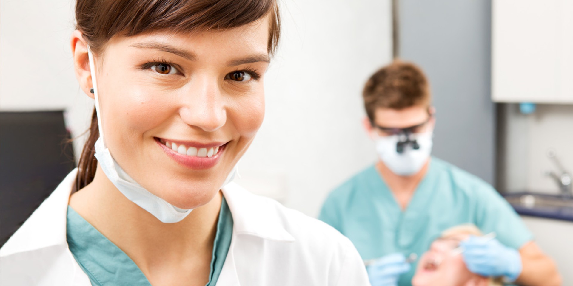 A woman in scrubs and a facemask under her chin is smiling while in the background a man is performing dental work on what appears to be a practice dummy