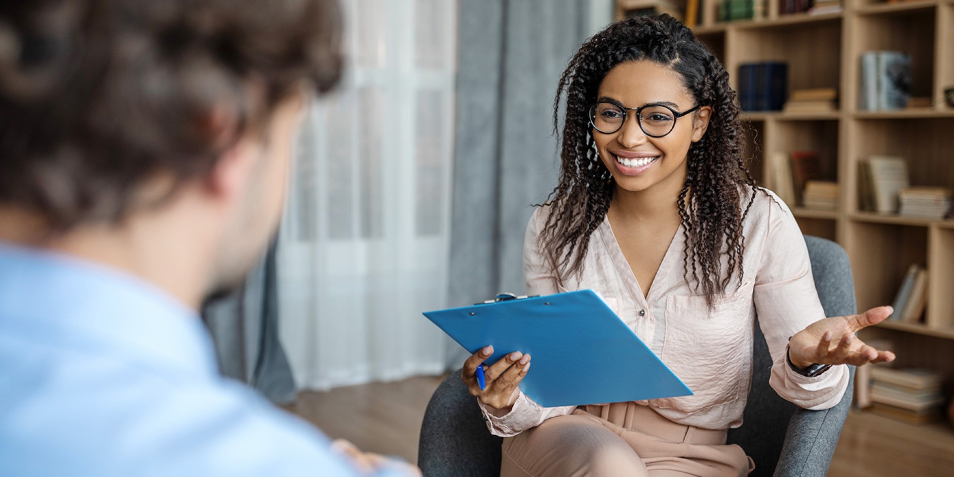 Woman sitting in chair smiling, holding a clip board, while giving life advice to someone in the foreground of the picture.